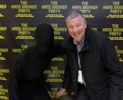 Photo of a man in front of a black wall with yellow text on it that says "The Hans Brinker Party"